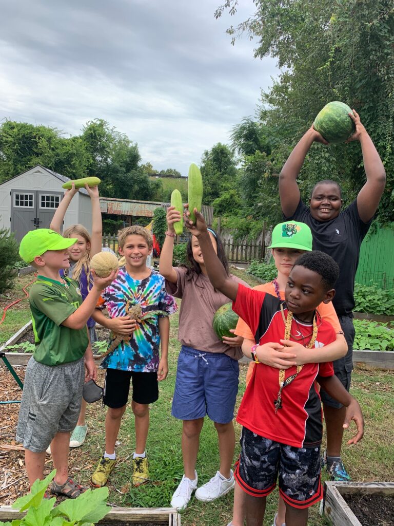 A group of joyous children stand in a school garden, holding up the melons and cucumbers they have just picked!